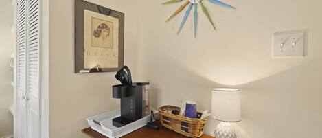 A room with a coffee maker on a wooden table, a framed picture on the wall, a star-shaped clock, and a lamp.