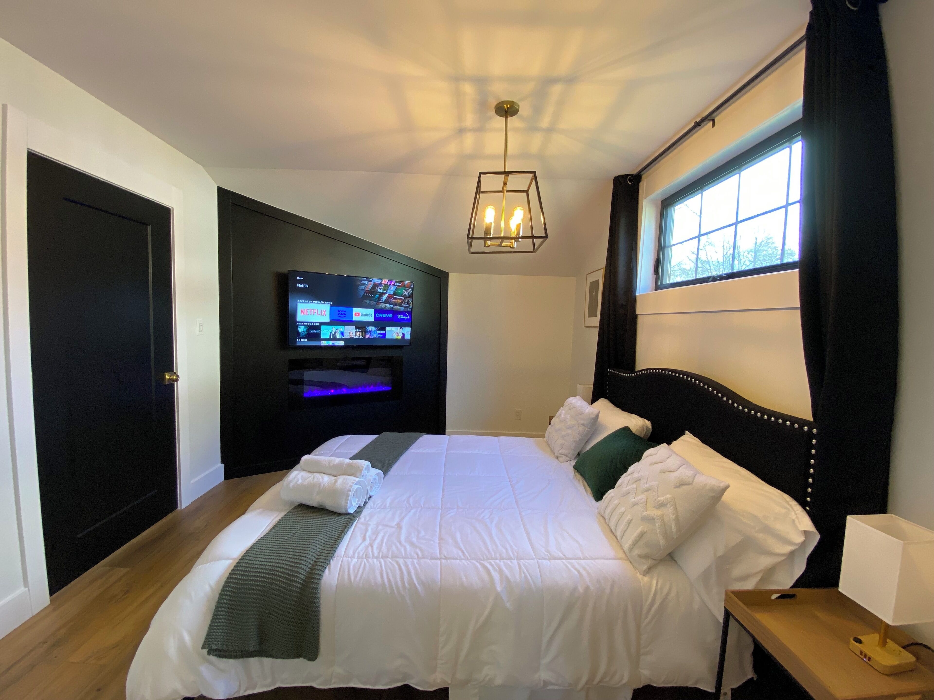 A hotel room with a bed, a TV mounted on the wall, a window with curtains, and a bedside table with a lamp.