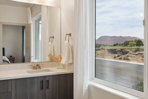 Un bagno con uno specchio grande, un lavandino e una finestra che si affaccia su una strada e montagne.