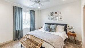 Ein Schlafzimmer mit einem Bett, einem Deckenventilator, einem Fenster mit Vorhängen, einem Nachttisch mit Lampe und Wandbildern.