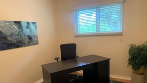 A room with a dark wooden desk, a black office chair, a blue rug, a window with blinds, and a potted plant. - Centrally located one bedroom + Office (Calgary)