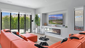 A modern living room with orange sofas, a fireplace, a large TV, and a view of greenery through glass doors. - New+Modern/Pool/Spa/Gameroom/Poker/Massage chair (Indio)
