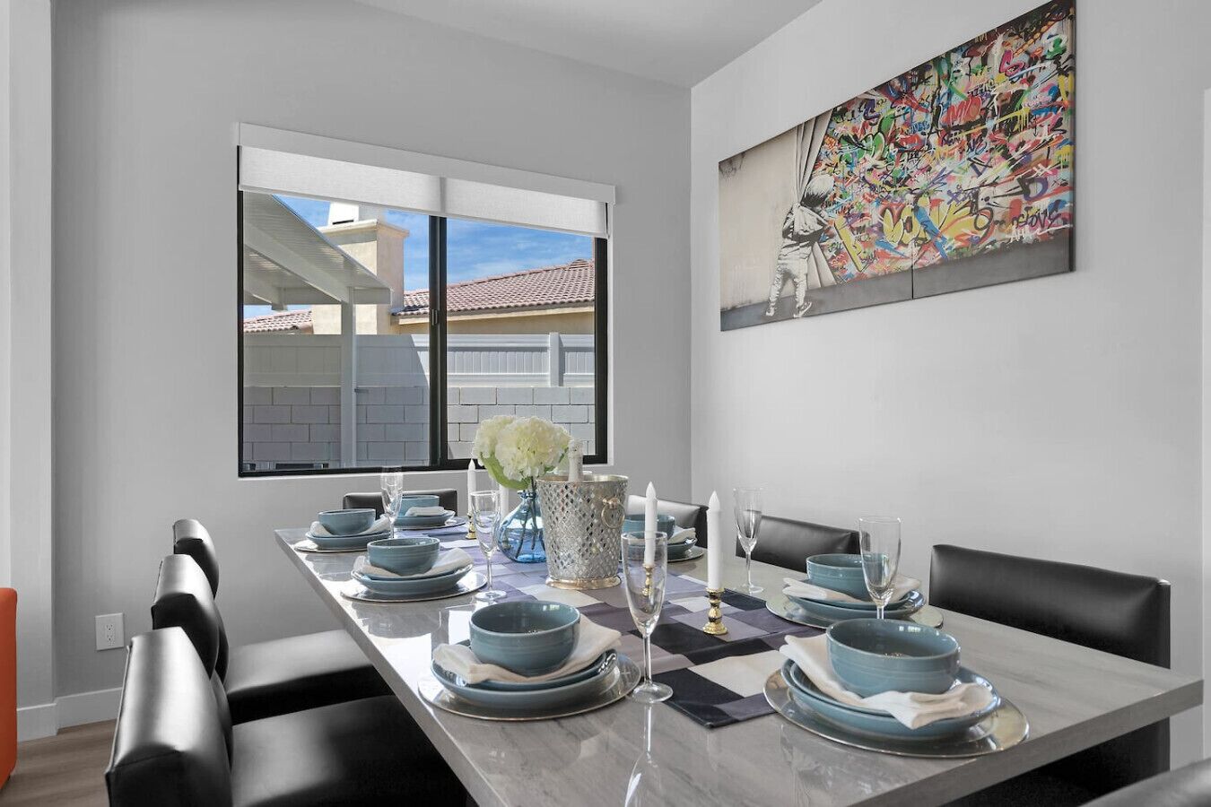 A modern dining room with a long table set for six, featuring blue and white plates, a vase with white flowers, and a colorful abstract painting on the wall.