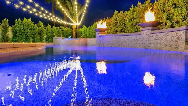 A swimming pool with string lights overhead, surrounded by greenery and a tiled wall. - New+Modern/Pool/Spa/Gameroom/Poker/Massage chair (Indio)