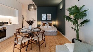 Une chambre d’hôtel compacte dotée d’une kitchenette, d’un coin repas avec une table dressée pour deux personnes, d’un lit avec une tête de lit sombre et d’une grande plante en pot.