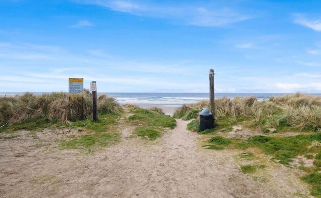 Un sentier de sable qui longe la plage et mène à l’océan, avec un panneau indicateur sur la gauche.