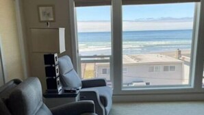 Una sala de estar con un ventanal grande que ofrece vista a la playa y al océano. Hay dos sofás grises y una mesita con una lámpara.