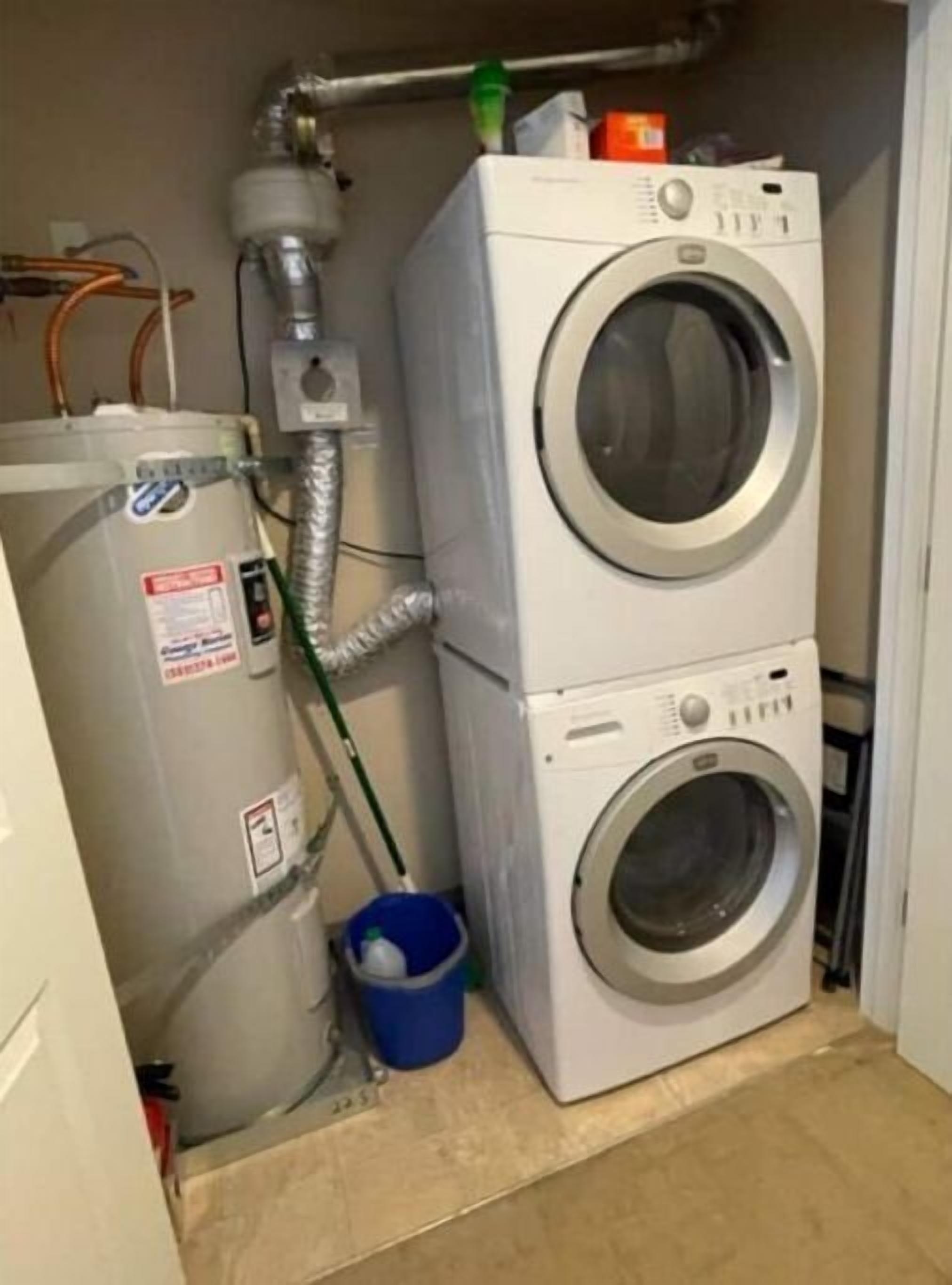 A compact laundry room with a stacked washer and dryer, a water heater, and a blue bucket.