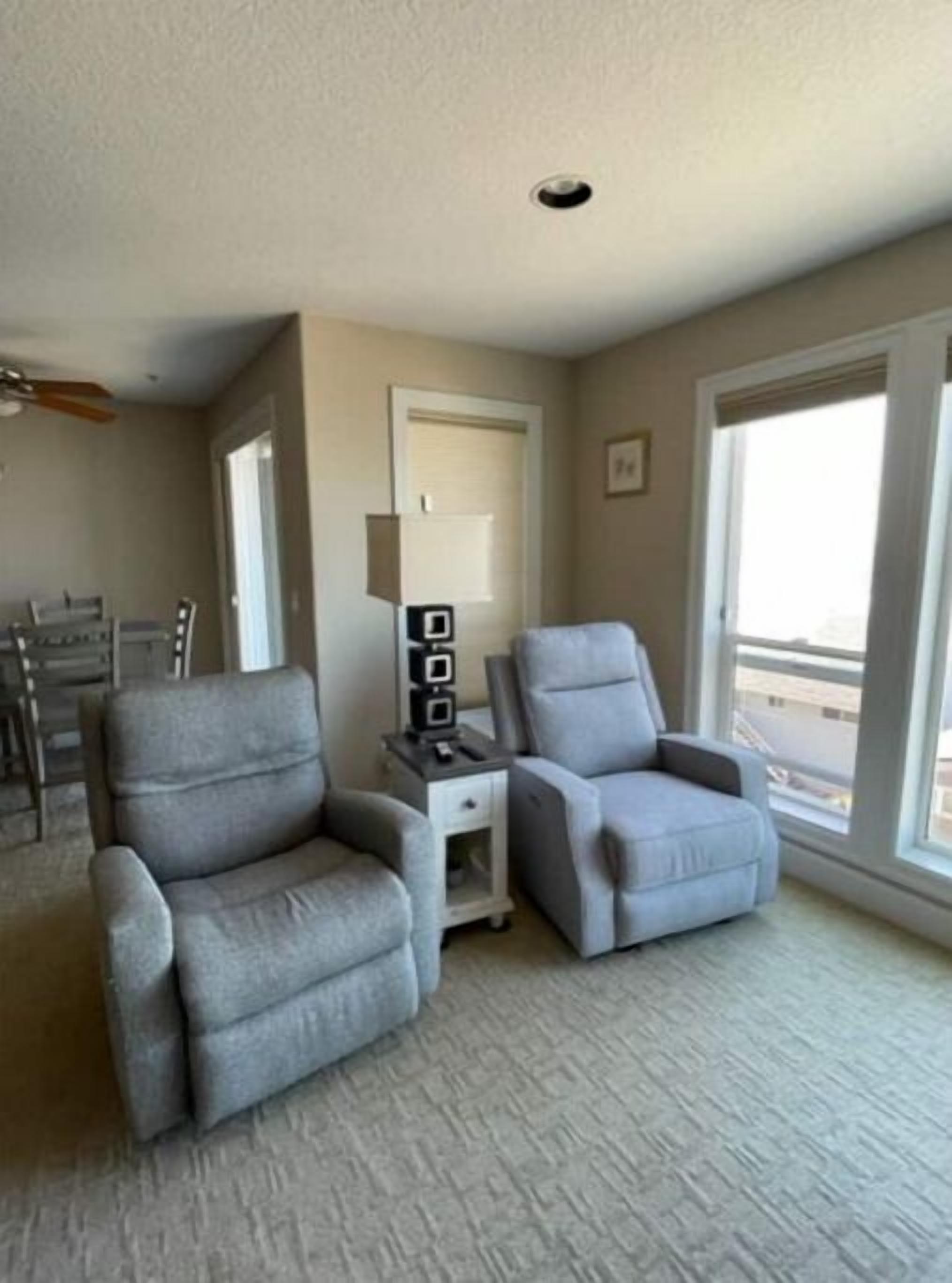 Un salon avec deux fauteuils gris, une petite table d’appoint blanche et un ventilateur de plafond.