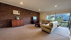A living room with a brick wall, a wooden cabinet, a leather sofa, a coffee table, and a television. - Hill top view house, high speed wifi, Netflix & Prime Video (Sunbury)