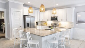 Una cocina moderna con armarios blancos, una isla de granito, taburetes de bar blancos e iluminación colgante.