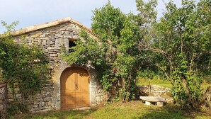 Ein rustikales Steinhaus mit einer Holztür, umgeben von Grünpflanzen und einer Steinsitzbank.