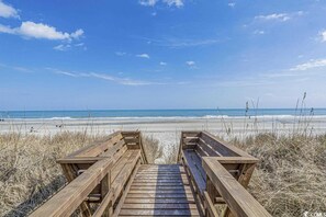 Une promenade en bois menant à une plage, avec un ciel dégagé et une vue sur l’océan.