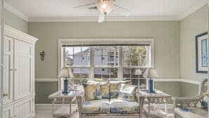 Um quarto com uma grande janela, um ventilador de teto, um sofá de vime e um armário branco.