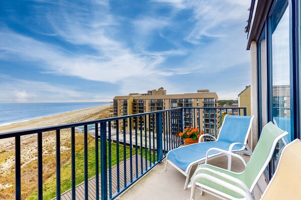 Enjoy stunning views of the beach from this balcony, featuring comfortable lounge chairs and a metal railing.