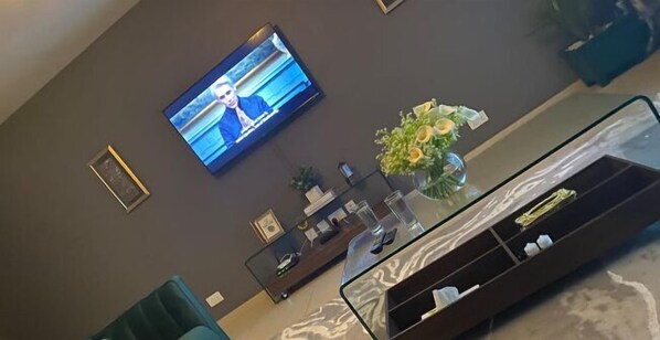 Een woonkamer met een flatscreen televisie aan de muur, een glazen salontafel en een decoratieve vaas.