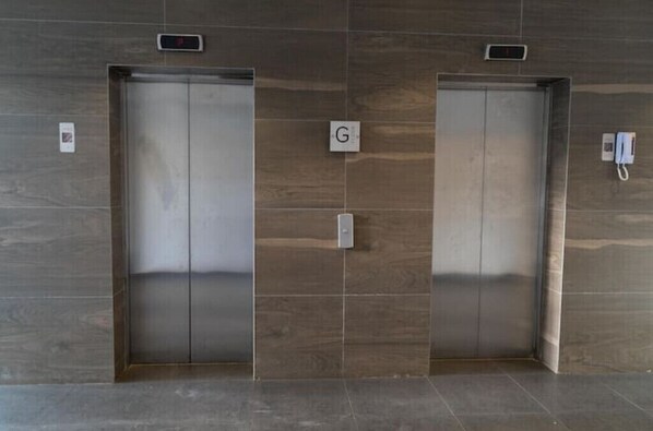 Two elevator doors with a digital display above each, a sign with the letter 'G' between them, and a phone mounted on the wall to the right.