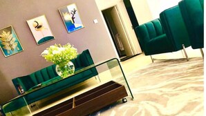 Um lobby moderno com um sofá verde, uma mesa de centro de vidro e um vaso de flores. Há três obras de arte emolduradas na parede e uma porta ao fundo.