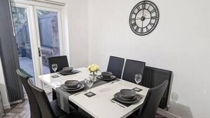 Un'area pranzo con tavolo bianco apparecchiato per quattro, sedie nere, un orologio appeso al muro e una finestra vista esterna.