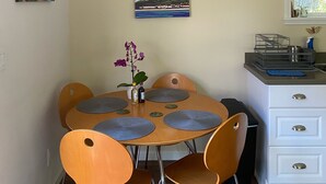 Uma sala de jantar com uma mesa redonda, quatro cadeiras e um vaso com flores. Há duas pinturas na parede, uma pia com janela e um armário branco com gavetas.