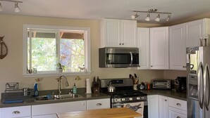 Cozinha com armários brancos, eletrodomésticos de aço inoxidável, mesa de madeira e janela com vista para área verde.