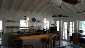 A spacious kitchen with a wooden island, bar stools, and a dining area with a table and chairs. There are ceiling fans, pendant lights, and a view through large windows.