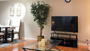 Ein Wohnzimmer mit Glastisch, einer Topfpflanze, einem Flachbildfernseher, einem Esstisch mit Stühlen und einer Uhr an der Wand.