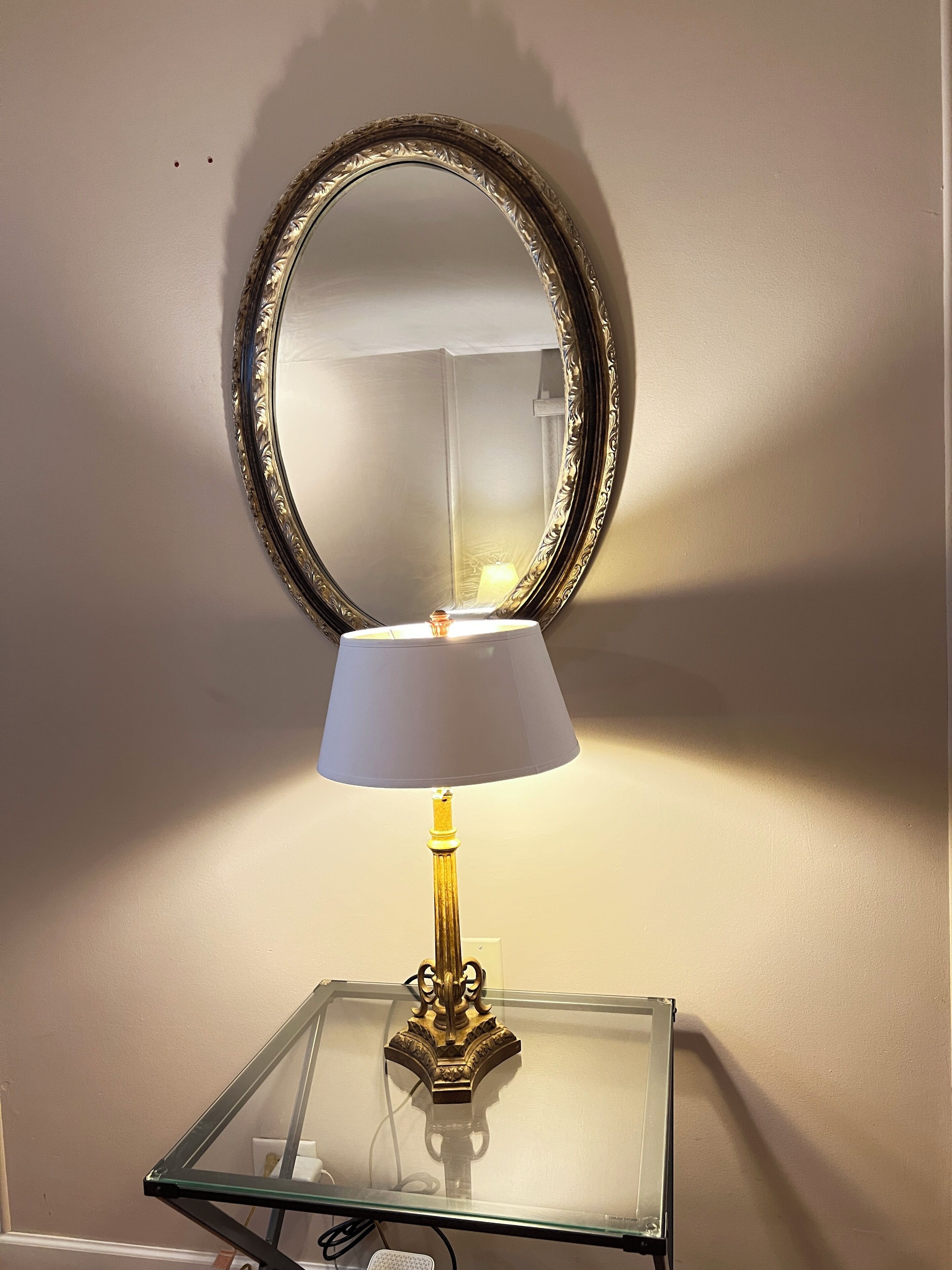 A table with a lamp and a mirror on a glass-top table against a wall.