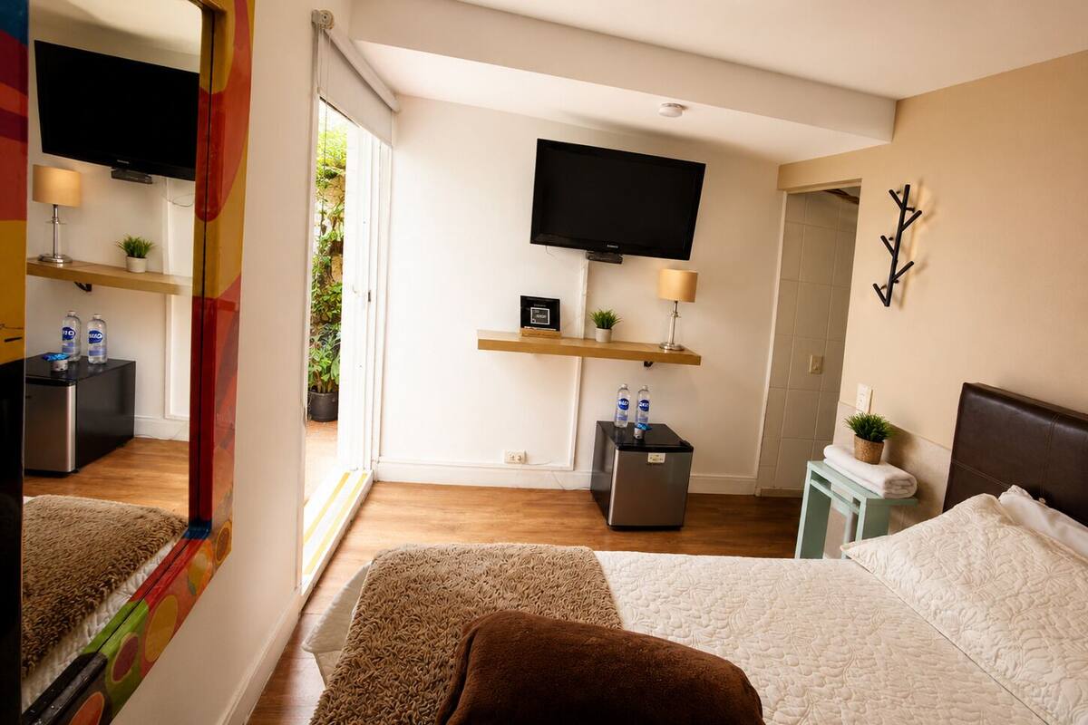 A bedroom with a bed, a TV, a small desk, a refrigerator, and a bathroom door.