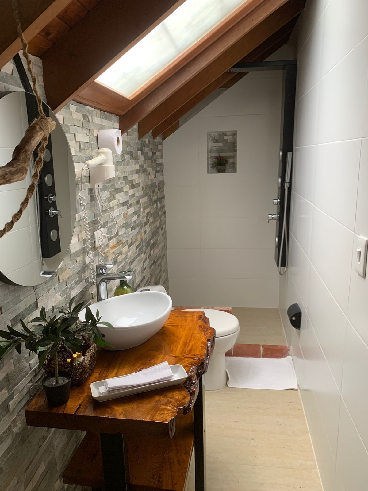 Ein modernes Badezimmer mit einer Wand aus Steinmosaik, einem Holzbadezimmermöbel, einem großen weißen Waschbecken, einer Toilette und einem Oberlicht.