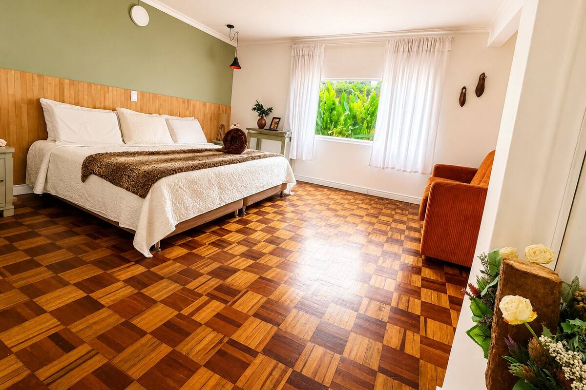 A bedroom with a wooden floor, a bed with white linens, a nightstand, a window with curtains, and a plant.