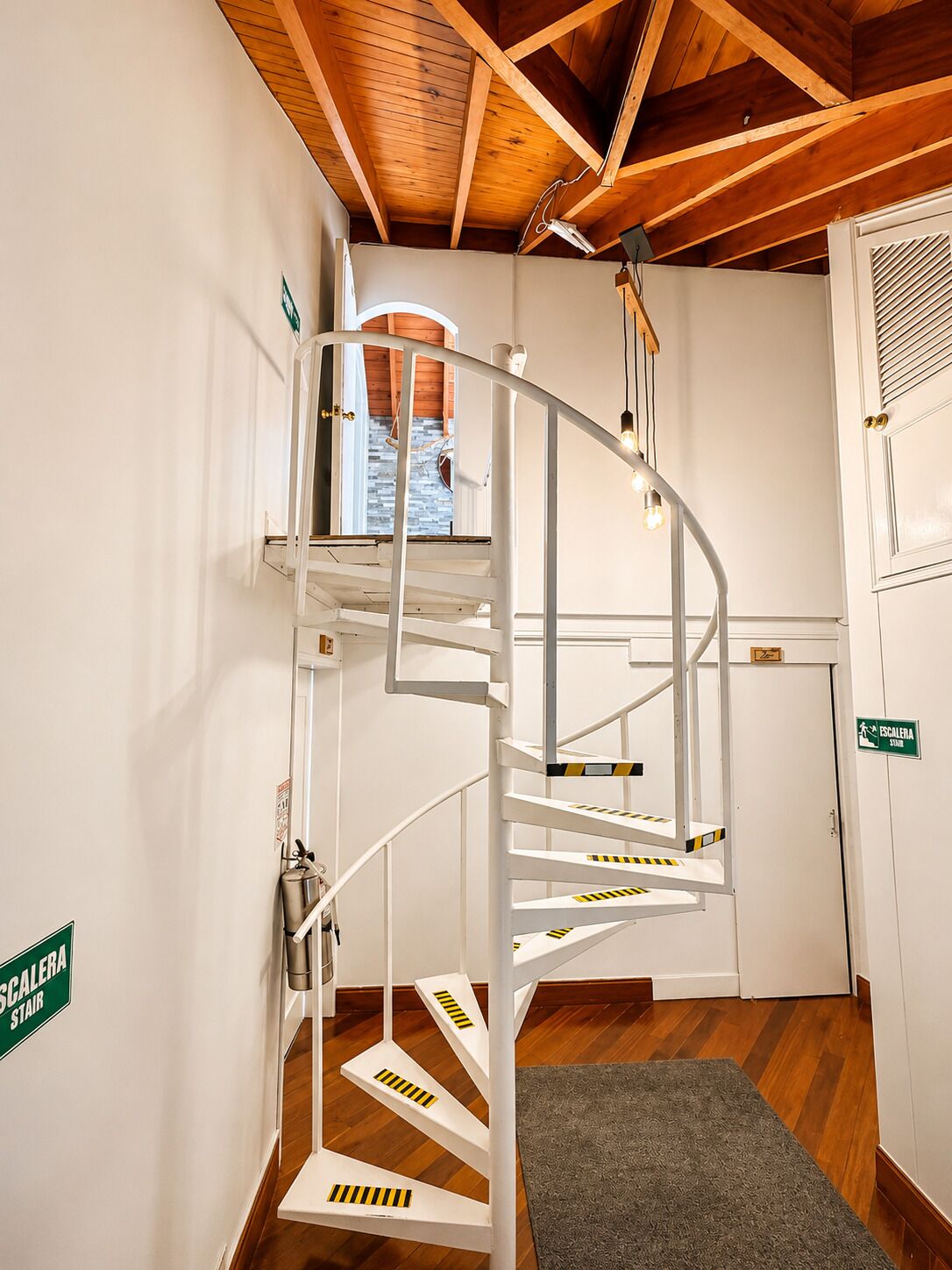 Un escalier en colimaçon avec une rampe en métal, un plafond en bois et une porte portant l’inscription « ESCALERA STAIR ».