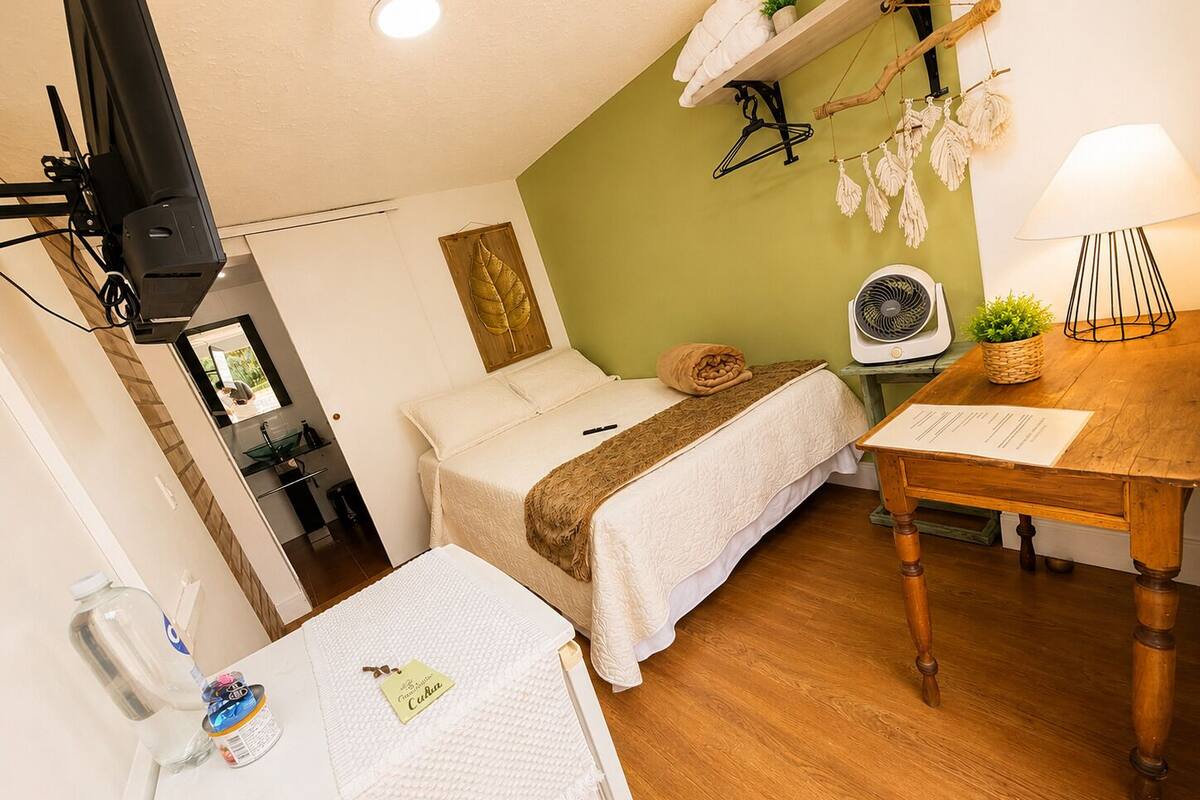 A bedroom with a bed, a bedside table with a lamp, a fan, and a small table with a menu.