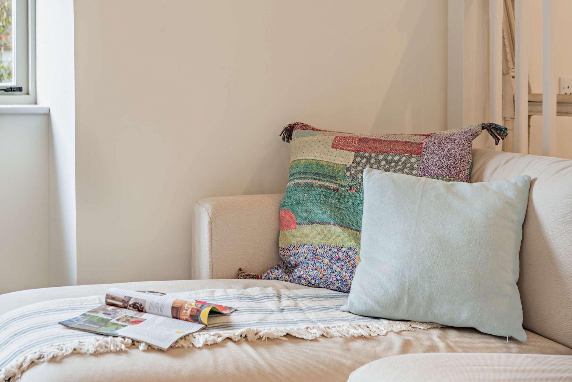 A cozy room with a beige sofa, a colorful patchwork pillow, and a magazine on the cushion.