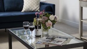 Een glazen salontafel met een fles wijn, twee wijnglazen en een vaas met bloemen.