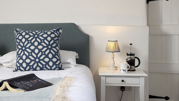Een keurig opgemaakt bed met een sierkussen, een bedlampje, een koffiezetapparaat en een boek op het nachtkastje.