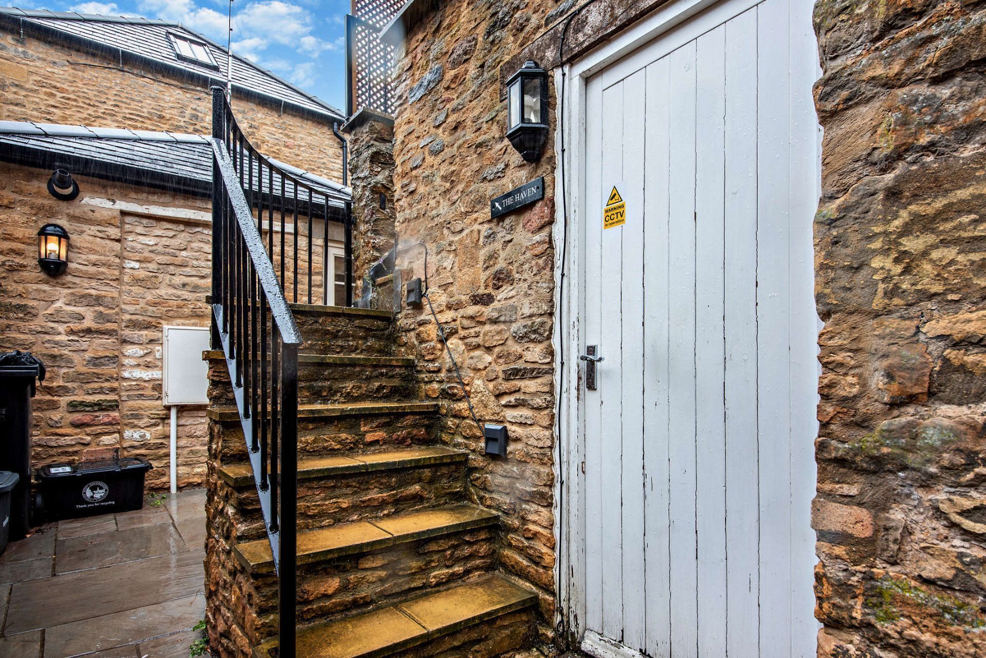 A stone staircase leading to a white door with a CCTV warning sign, a lantern, and a trash bin labeled 'Thank you for recycling.'