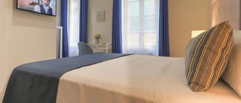 Une chambre d’hôtel avec un lit, un téléviseur fixé au mur, un bureau et une fenêtre avec des rideaux.