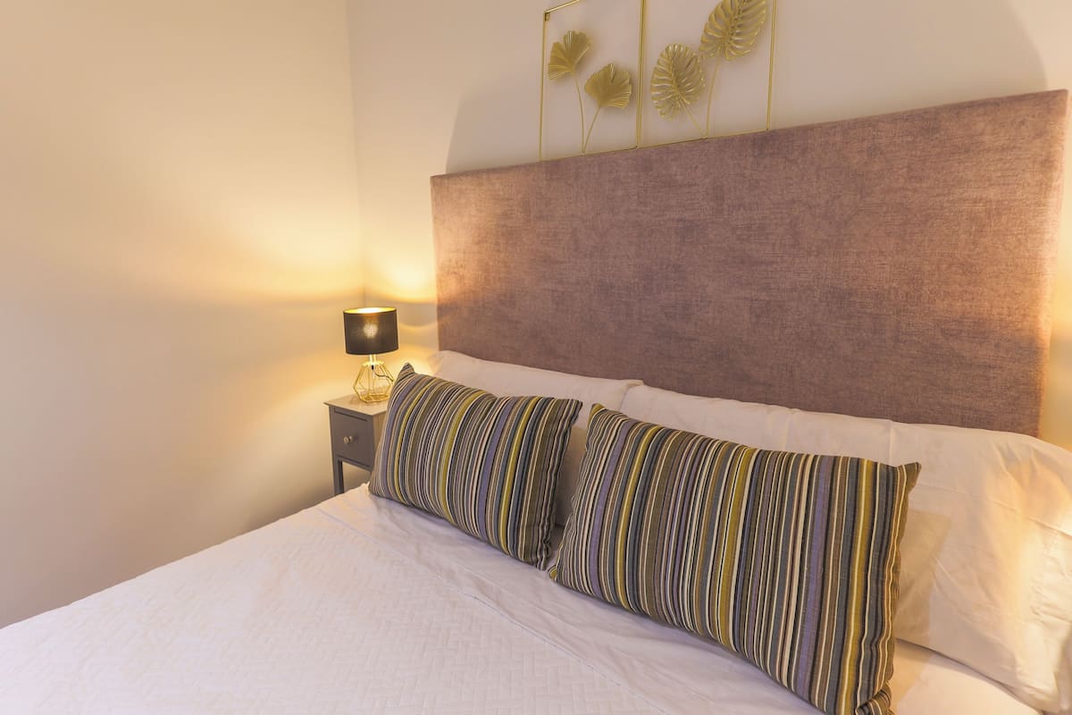 Ein ordentlich bezogenes Bett mit gestreiften Kissen, einer Nachttischlampe und einem Wandbild mit Blattformen.
