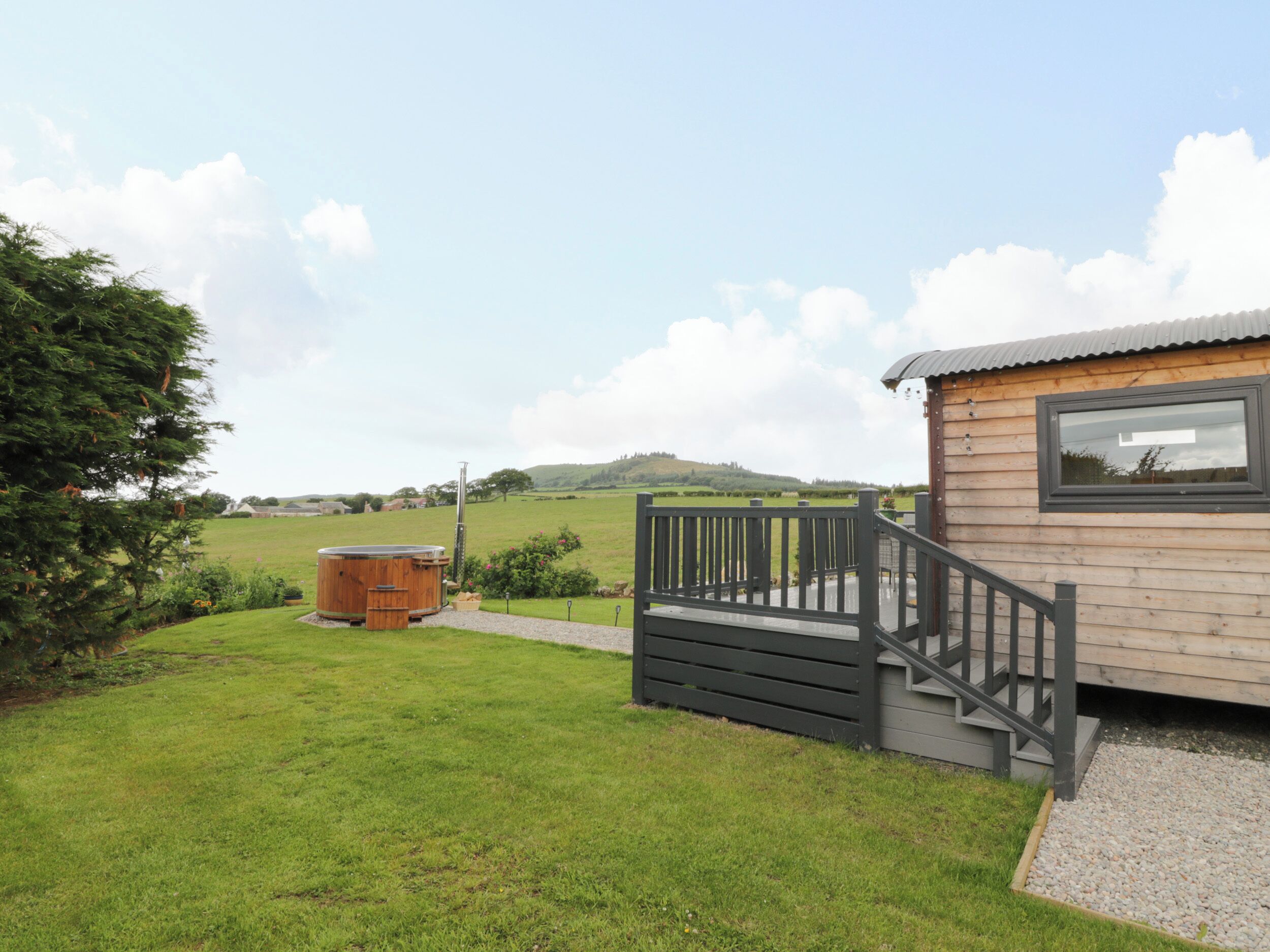 A wooden cabin with a staircase leading to a deck, surrounded by a grassy yard and a hot tub.