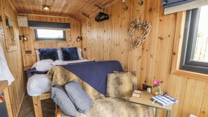 Interior acogedor de una cabaña con paredes de madera, una cama con ropa de cama azul, un banco cubierto de pieles y una mesita con un libro y flores.