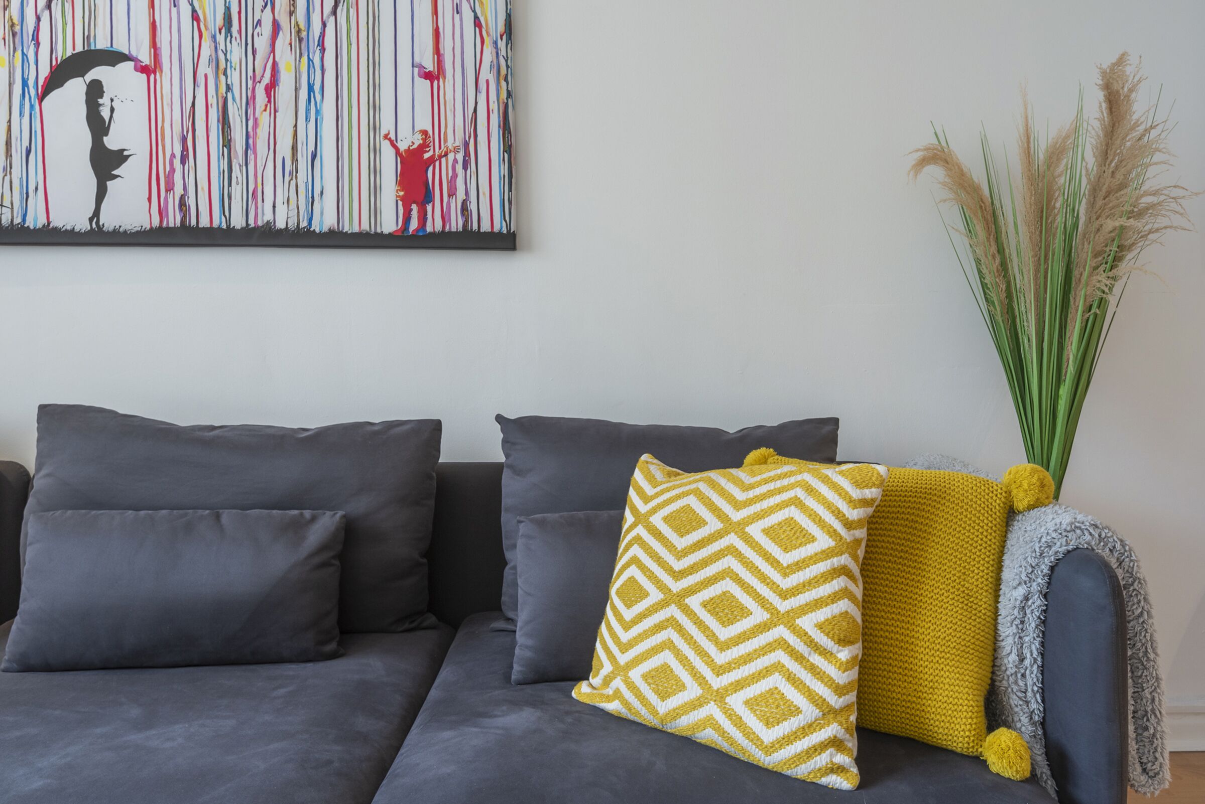 Un canapé gris avec trois coussins, un oreiller jaune à motifs et une plante décorative aux grandes feuilles vertes.