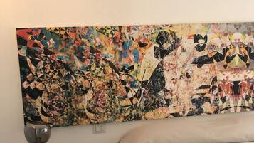 Une chambre avec un grand tableau abstrait au mur, un lit recouvert d’une couverture blanche, une petite table de chevet en bois avec une lampe, et un sol carrelé à motifs hexagonaux.