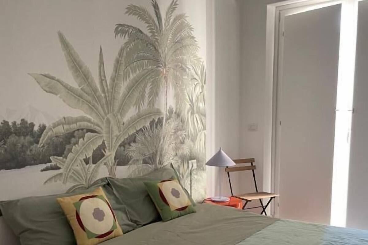 Un dormitorio con cama, almohadas, mesita de noche, lámpara y un mural de palmeras.
