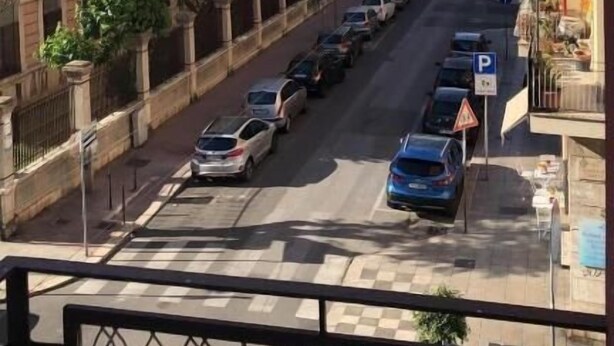Vista da rua com carros estacionados, faixa de pedestres e edifícios com varandas.