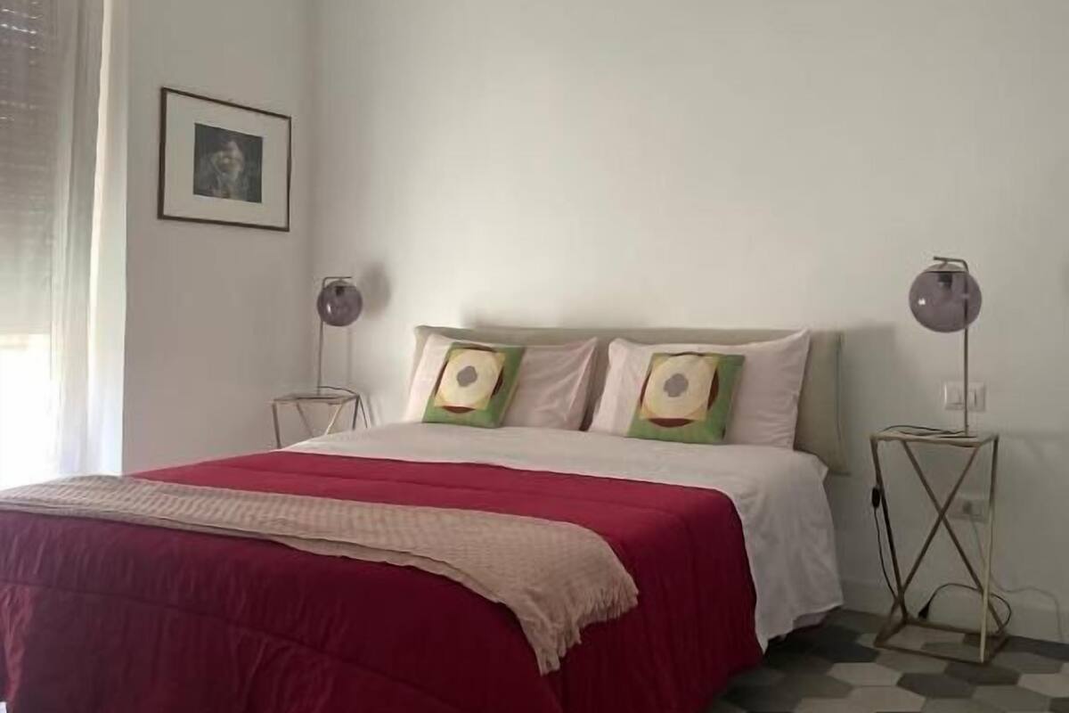 Un dormitorio con cama, lámpara de noche, un cuadro en la pared y ventana con persianas.
