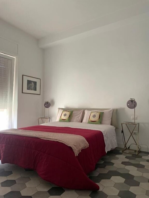 A bedroom with a bed, a bedside lamp, a picture on the wall, and a window with blinds.