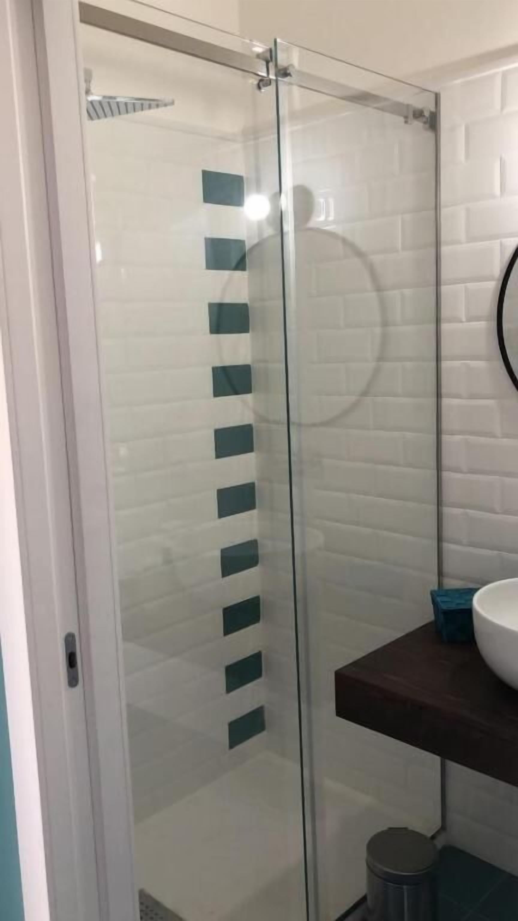 A glass-enclosed shower with a white tiled wall, a wooden shelf with a sink, and a trash bin.