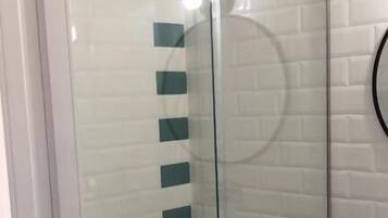 A glass-enclosed shower with a white tiled wall, a wooden shelf with a sink, and a trash bin.