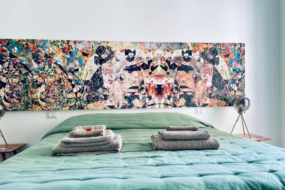 Un dormitorio con un cuadro abstracto grande en la pared, un edredón verde y dos toallas dobladas sobre la cama.